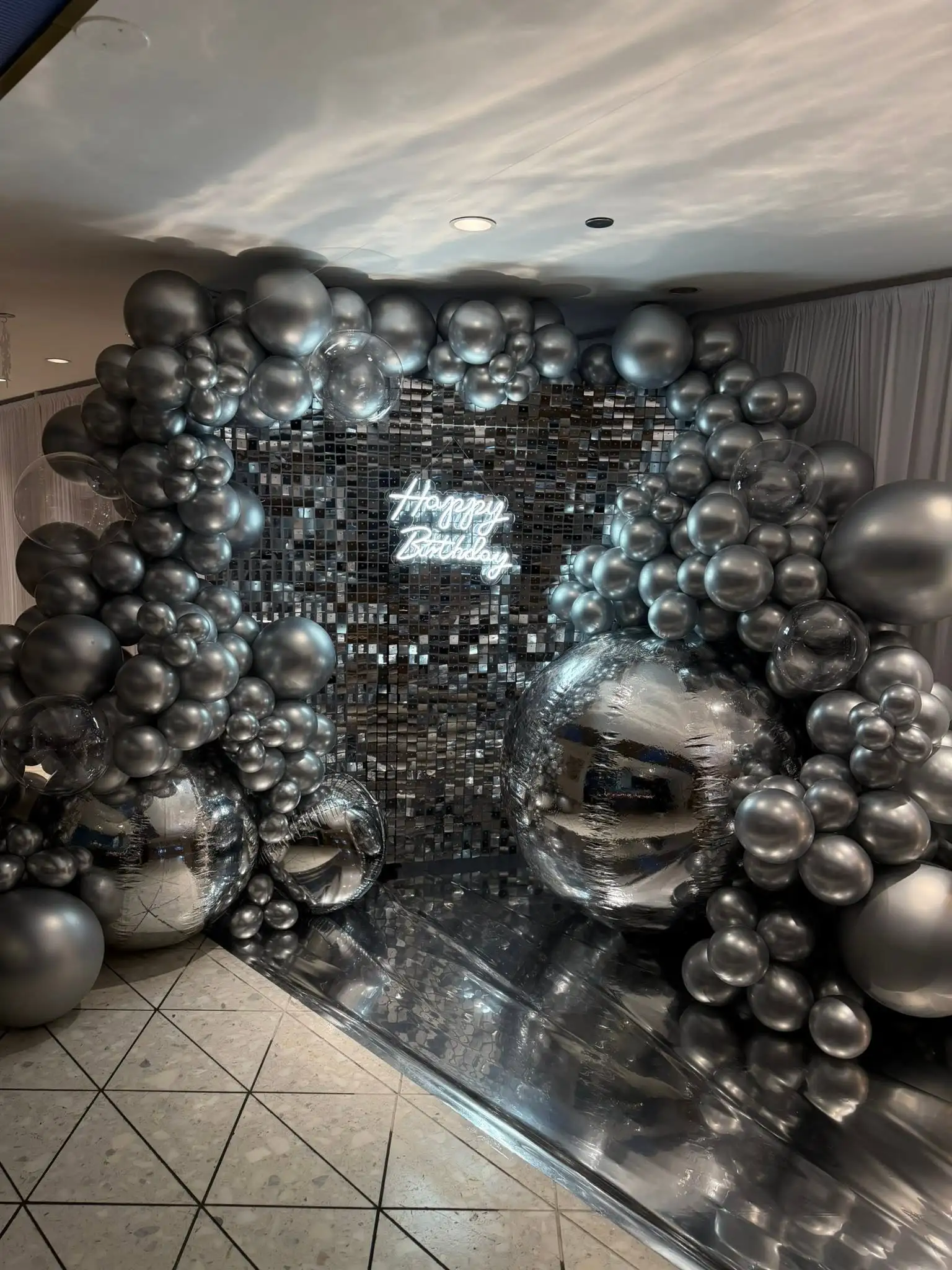 Silver chrome balloon arch and mirror shimmer wall with a Happy Birthday neon sign
